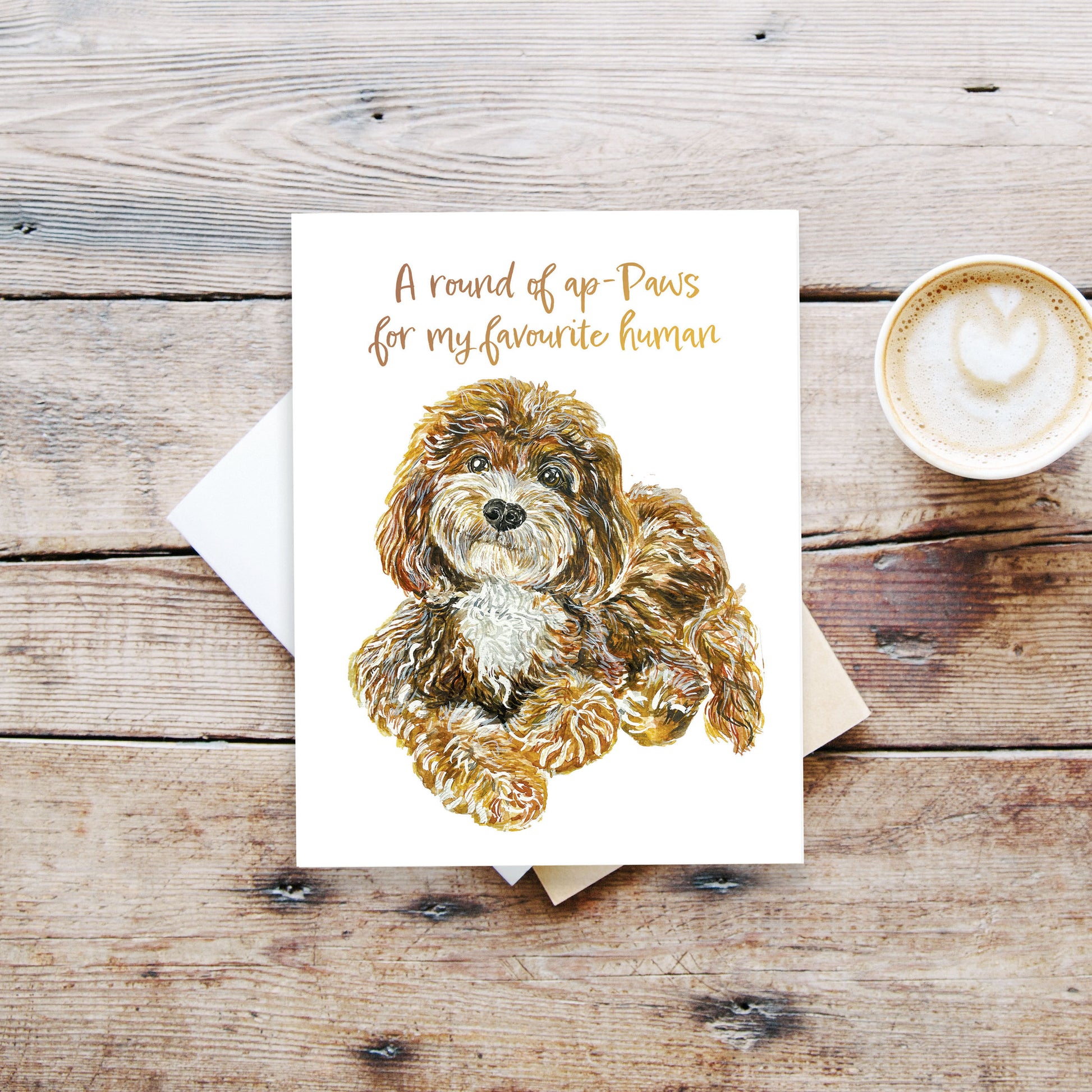 A greeting card and a cup of latte on a table. The card has a golden doodle looking sweetly at you. The text reads: A round of "ap-paws" for my favourite human. (Play on word "Applause")